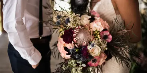 A bride in a white lace dress holding a large, lush wedding bouquet with pink and white flowers and dark greenery.