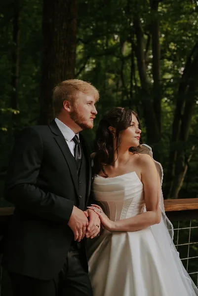 A bride and groom standing together in a forest setting, holding hands.