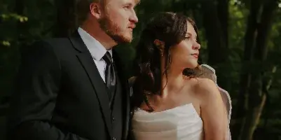 A bride and groom standing together in a forest setting, holding hands.
