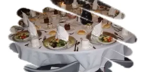 A round catering table set with white linens, folded napkins, plates of salad and rolls, and formal place settings.