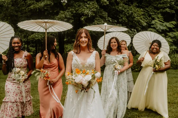 A bride and her bridesmaids standing outdoors in a garden, holding white parasols and bouquets.