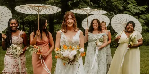 A bride and her bridesmaids standing outdoors in a garden, holding white parasols and bouquets.
