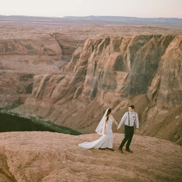 A bride and groom walking hand-in-hand on a rocky cliff edge overlooking a vast canyon landscape.