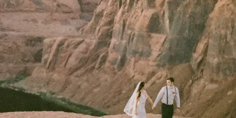 A bride and groom walking hand-in-hand on a rocky cliff edge overlooking a vast canyon landscape.