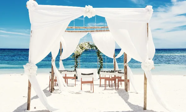 A white wedding canopy and floral arch set up on a sandy beach with the ocean in the background.