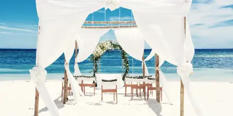 A white wedding canopy and floral arch set up on a sandy beach with the ocean in the background.