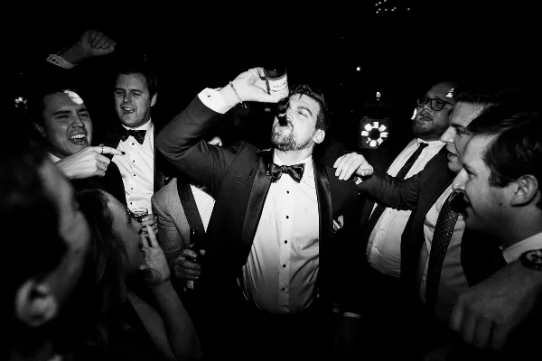 A black and white candid photo of a groom drinking from a bottle surrounded by his groomsmen at a wedding reception.