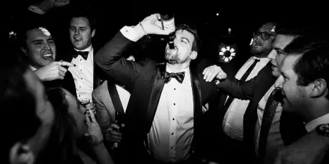A black and white candid photo of a groom drinking from a bottle surrounded by his groomsmen at a wedding reception.
