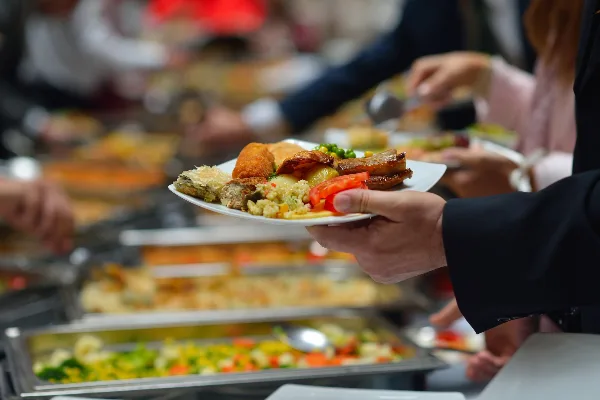 A server in a black suit holds a white plate filled with various food items, including meat, vegetables, and a fried item, in front of a blurred catering buffet line.