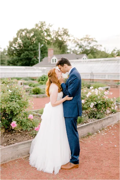 A bride and groom standing close together in a garden with rose bushes, the groom gently touching the bride's face.