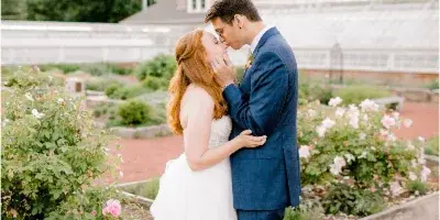 A bride and groom standing close together in a garden with rose bushes, the groom gently touching the bride's face.
