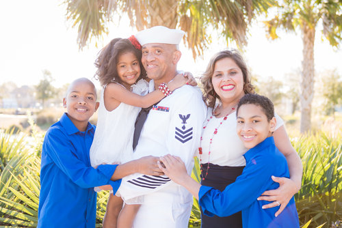 A family portrait featuring a man in a U.S. Navy uniform holding a young girl, with a woman and two boys in blue shirts, photographed outdoors with palm trees in the background.