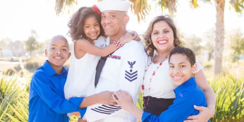 A family portrait featuring a man in a U.S. Navy uniform holding a young girl, with a woman and two boys in blue shirts, photographed outdoors with palm trees in the background.