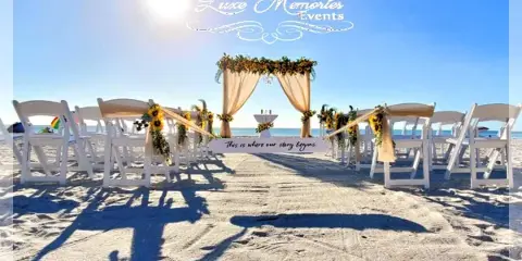 A beach wedding ceremony setup featuring a floral arch, draped fabric, and rows of white chairs.