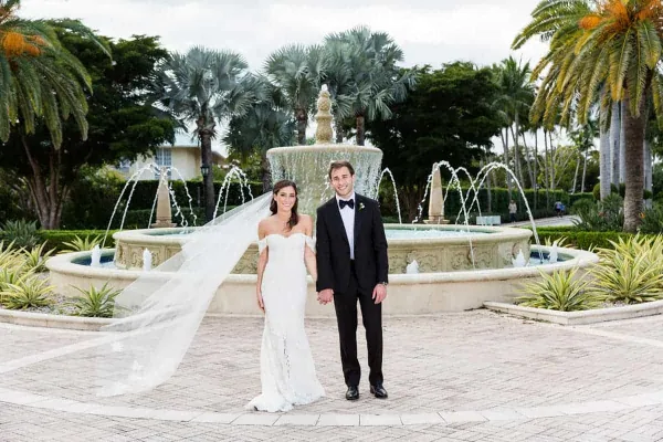 Bride and groom in formal wedding attire standing in front of a large ornate fountain with palm trees in the background.