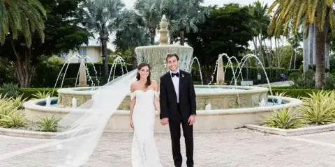 Bride and groom in formal wedding attire standing in front of a large ornate fountain with palm trees in the background.