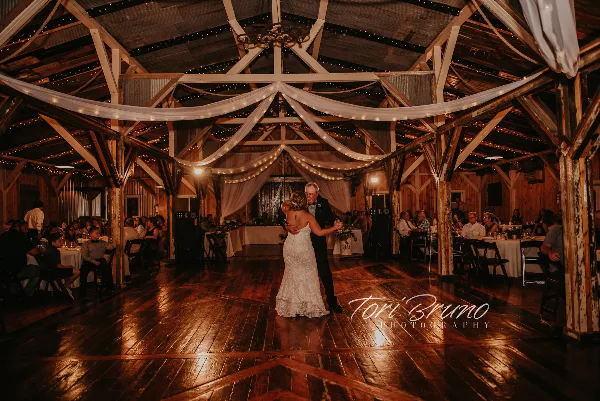 A bride and her father dancing in a rustic wooden barn reception hall with string lights and white drapery.