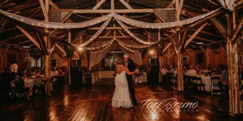 A bride and her father dancing in a rustic wooden barn reception hall with string lights and white drapery.