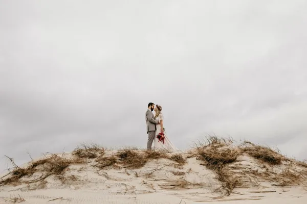 A bride and groom standing on a sand dune in a coastal environment.