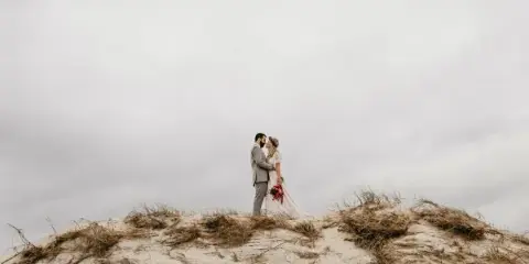 A bride and groom standing on a sand dune in a coastal environment.