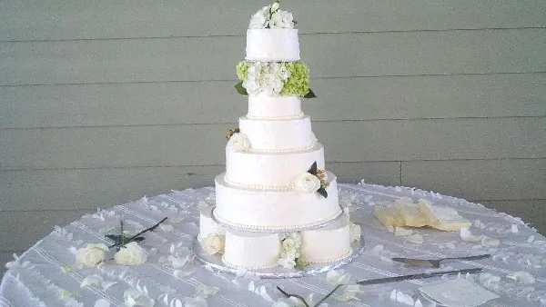 A five-tier white wedding cake decorated with white and green flowers, sitting on a table with a white tablecloth and scattered petals.