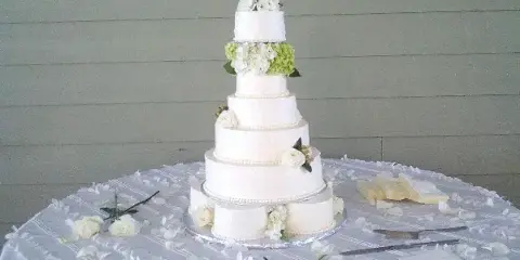 A five-tier white wedding cake decorated with white and green flowers, sitting on a table with a white tablecloth and scattered petals.