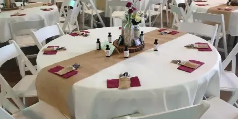 Interior of a wedding venue with round tables set with white linens, burlap runners, and floral centerpieces.