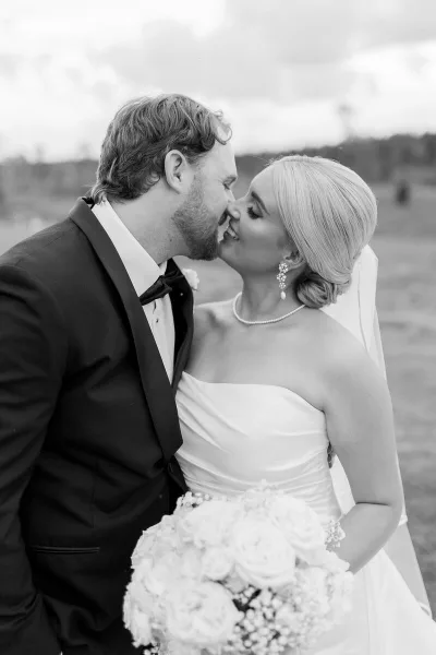A black and white photograph of a bride and groom kissing outdoors, with the bride holding a bouquet of white roses.
