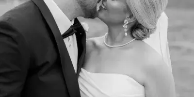 A black and white photograph of a bride and groom kissing outdoors, with the bride holding a bouquet of white roses.