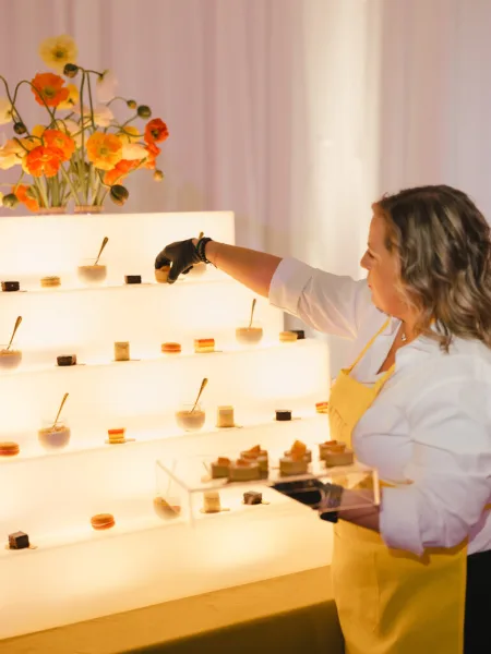 A catering staff member in a yellow apron arranges small desserts on an illuminated tiered display stand.