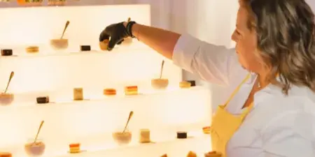 A catering staff member in a yellow apron arranges small desserts on an illuminated tiered display stand.