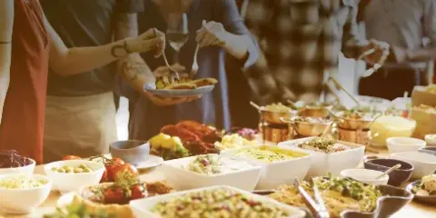 A catering buffet table filled with various dishes, salads, and bowls, with people serving themselves.
