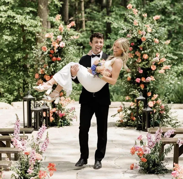 A groom carrying his bride during an outdoor wedding ceremony with floral decorations.