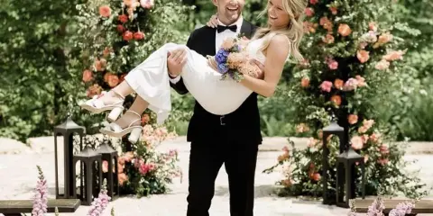 A groom carrying his bride during an outdoor wedding ceremony with floral decorations.