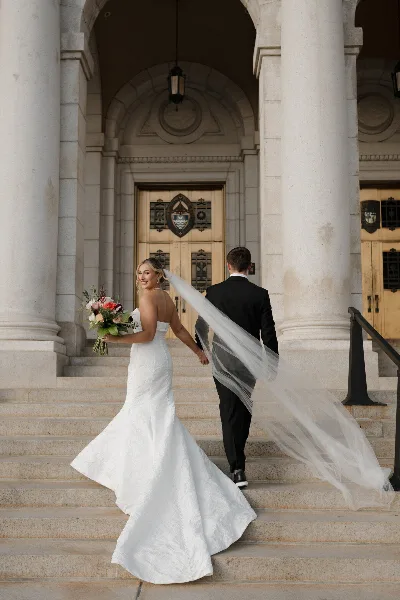 A bride in a white gown and a groom in a black suit walking up stone steps together, holding hands.