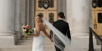 A bride in a white gown and a groom in a black suit walking up stone steps together, holding hands.