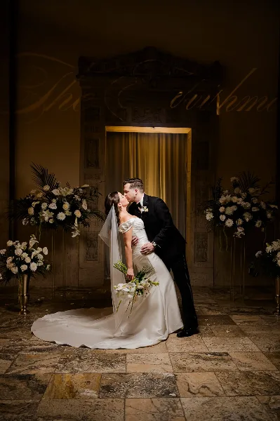 A bride and groom share a kiss in an elegant, dimly lit venue with floral arrangements.
