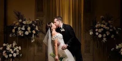 A bride and groom share a kiss in an elegant, dimly lit venue with floral arrangements.
