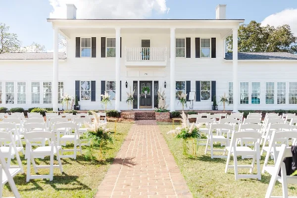 Outdoor wedding ceremony setup with white chairs and floral arrangements in front of a white farmhouse venue.