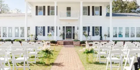Outdoor wedding ceremony setup with white chairs and floral arrangements in front of a white farmhouse venue.