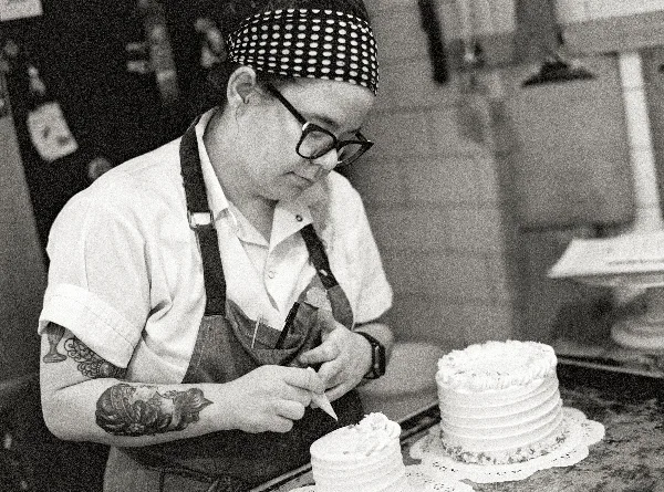 A baker wearing a polka-dot bandana and apron is piping white frosting onto a tiered cake in a bakery kitchen.