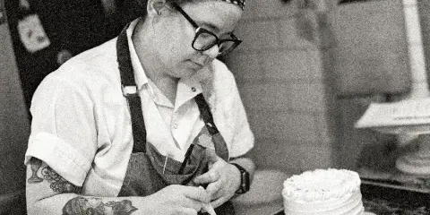 A baker wearing a polka-dot bandana and apron is piping white frosting onto a tiered cake in a bakery kitchen.