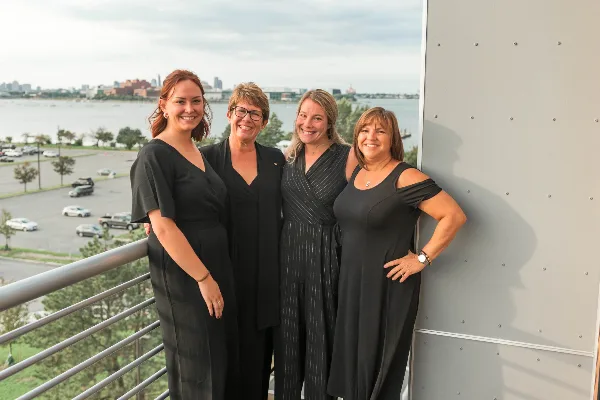 Four women in black professional attire standing together on a balcony overlooking a city and water.