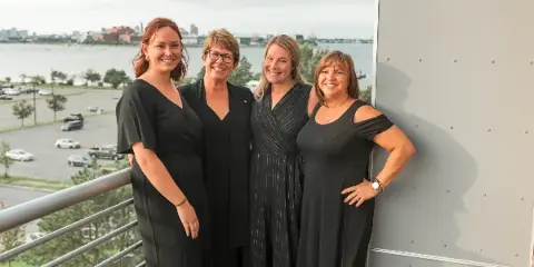 Four women in black professional attire standing together on a balcony overlooking a city and water.