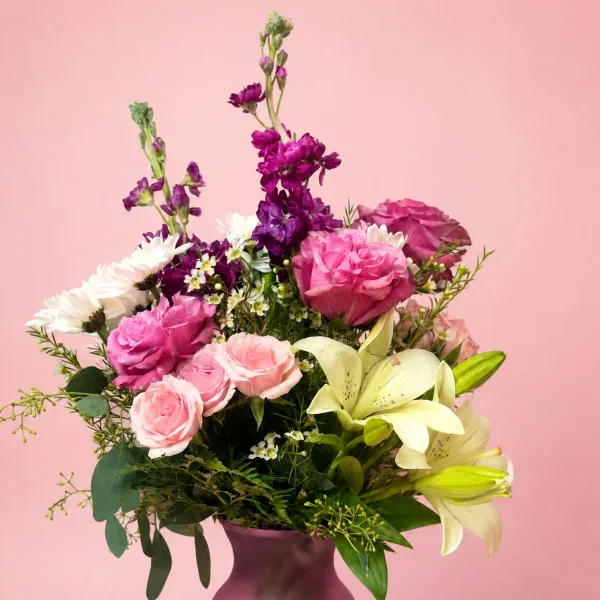 A floral arrangement featuring pink roses, white lilies, and purple stock in a purple vase against a pink background.
