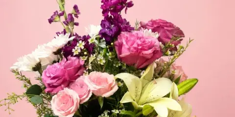 A floral arrangement featuring pink roses, white lilies, and purple stock in a purple vase against a pink background.