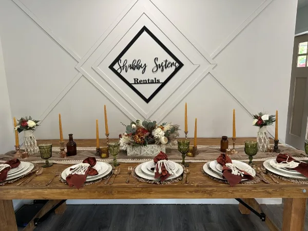 A rustic wooden wedding table set with white plates, burgundy napkins, green glassware, and floral centerpieces in front of a Shabby Sisters Rentals logo.
