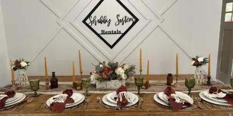 A rustic wooden wedding table set with white plates, burgundy napkins, green glassware, and floral centerpieces in front of a Shabby Sisters Rentals logo.