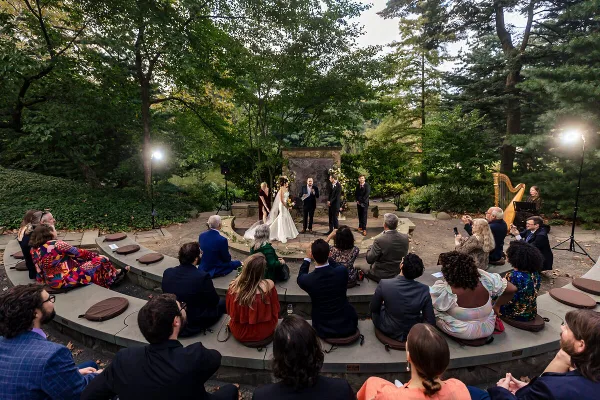 Outdoor wedding ceremony at a garden venue with guests seated in a circle around the couple and officiant.