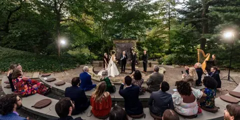 Outdoor wedding ceremony at a garden venue with guests seated in a circle around the couple and officiant.